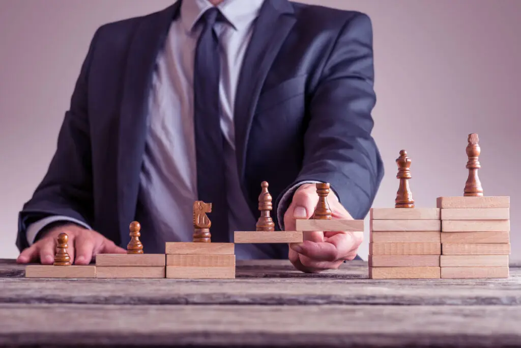 Homem de terno organizando peças de xadrez em blocos de madeira em diferentes alturas, simbolizando o planejamento estratégico em gestão de relacionamento com o cliente. A imagem representa conceitos de CRM (Customer Relationship Management) e desenvolvimento progressivo para aprimorar a experiência do cliente