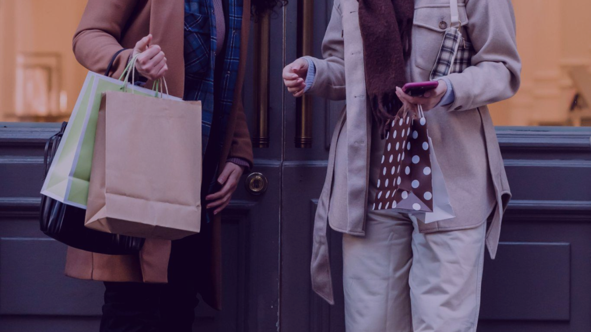 Duas mulheres com sacolas de compra e celular na mão, conversando sobre compras.