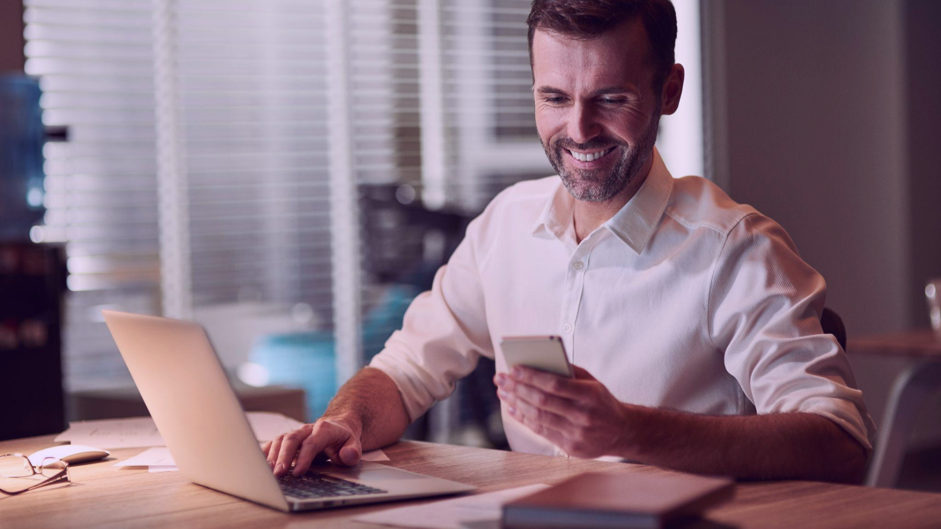 Homem sorridente sentado à mesa de trabalho, verificando o celular enquanto digita em um laptop, refletindo a eficácia do uso de estratégias omnichannel no varejo para impulsionar as vendas físicas.
